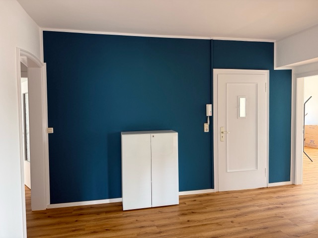 Flur mit petrolblauer Wand, weißer Tür mit kleinem Fenster und einem weißen Schrank auf hellem Holzboden