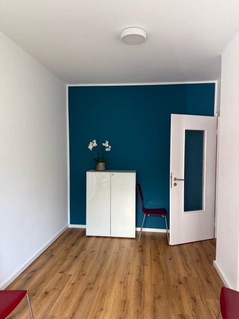 Kleiner Raum mit petrolblauer Wand, weißem Schrank mit weißer Orchidee, rotem Stuhl und angelehnter Tür