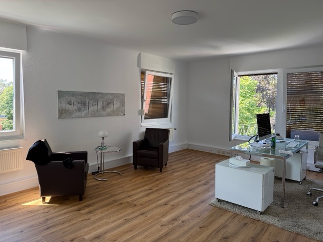 Heller Praxisraum mit Holzboden: zwei dunkle Ledersessel, ein kleiner Glastisch mit Leuchte, ein Bild an der Wand und ein Schreibtisch vor Fenstern mit Blick in grüne Bäume
